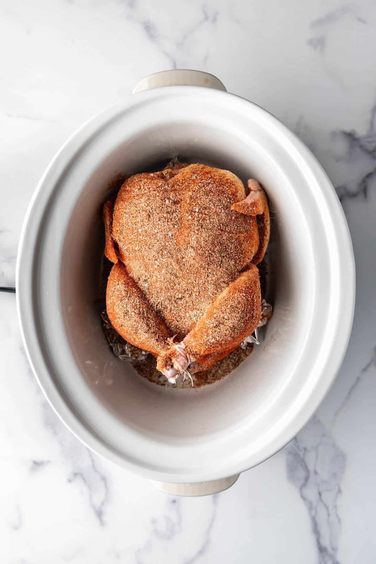 An overhead image of a chicken rubbed with seasoning in a white slow cooker.