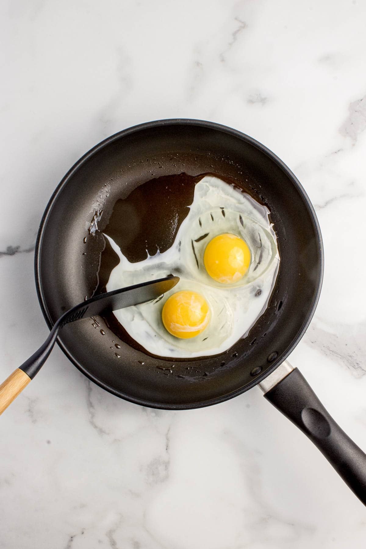 A spatula being used to "break" the egg whites on two eggs being fried in a pan.