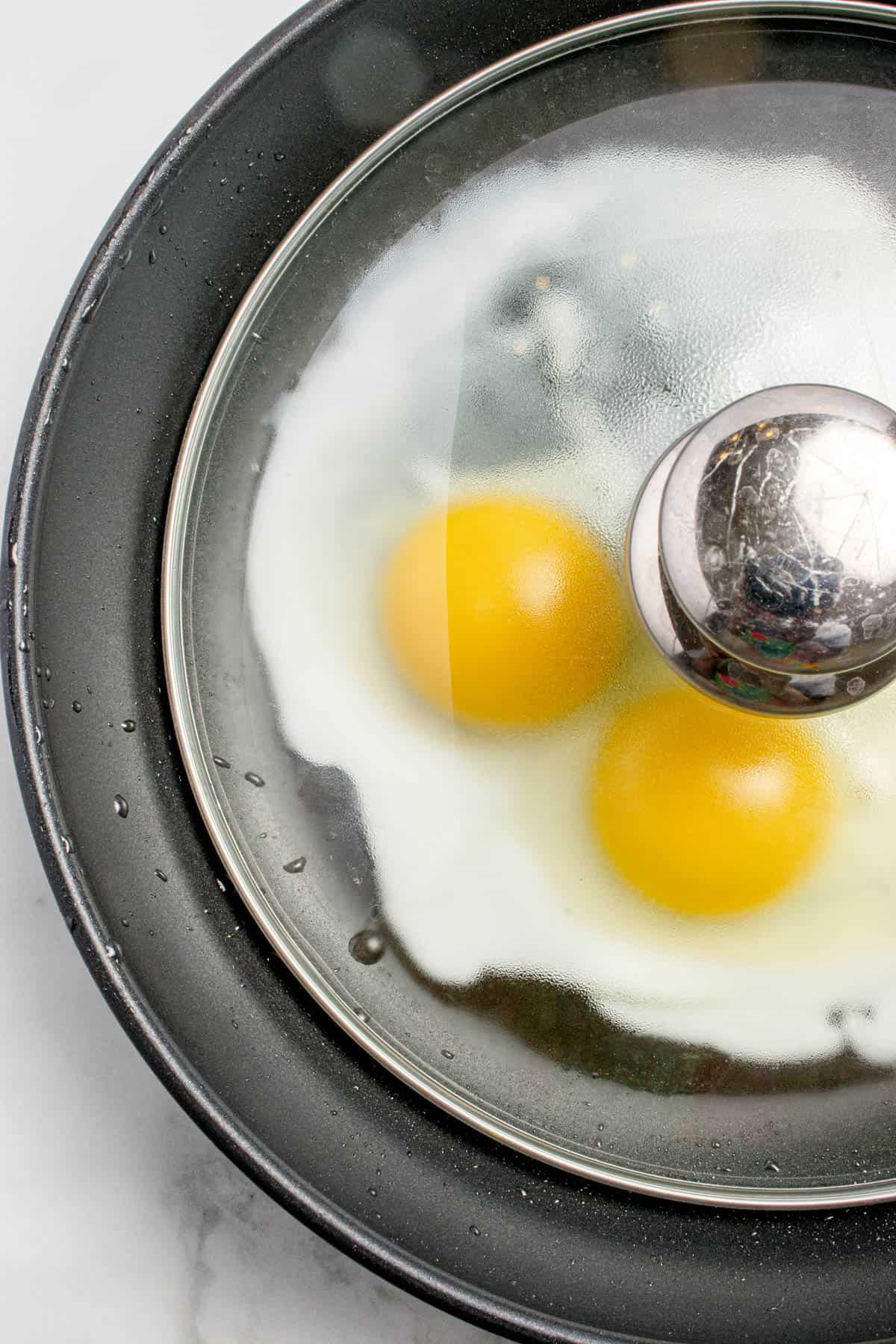 A close up image of a glass lid over two fried eggs cooking sunny side up in a frying pan.