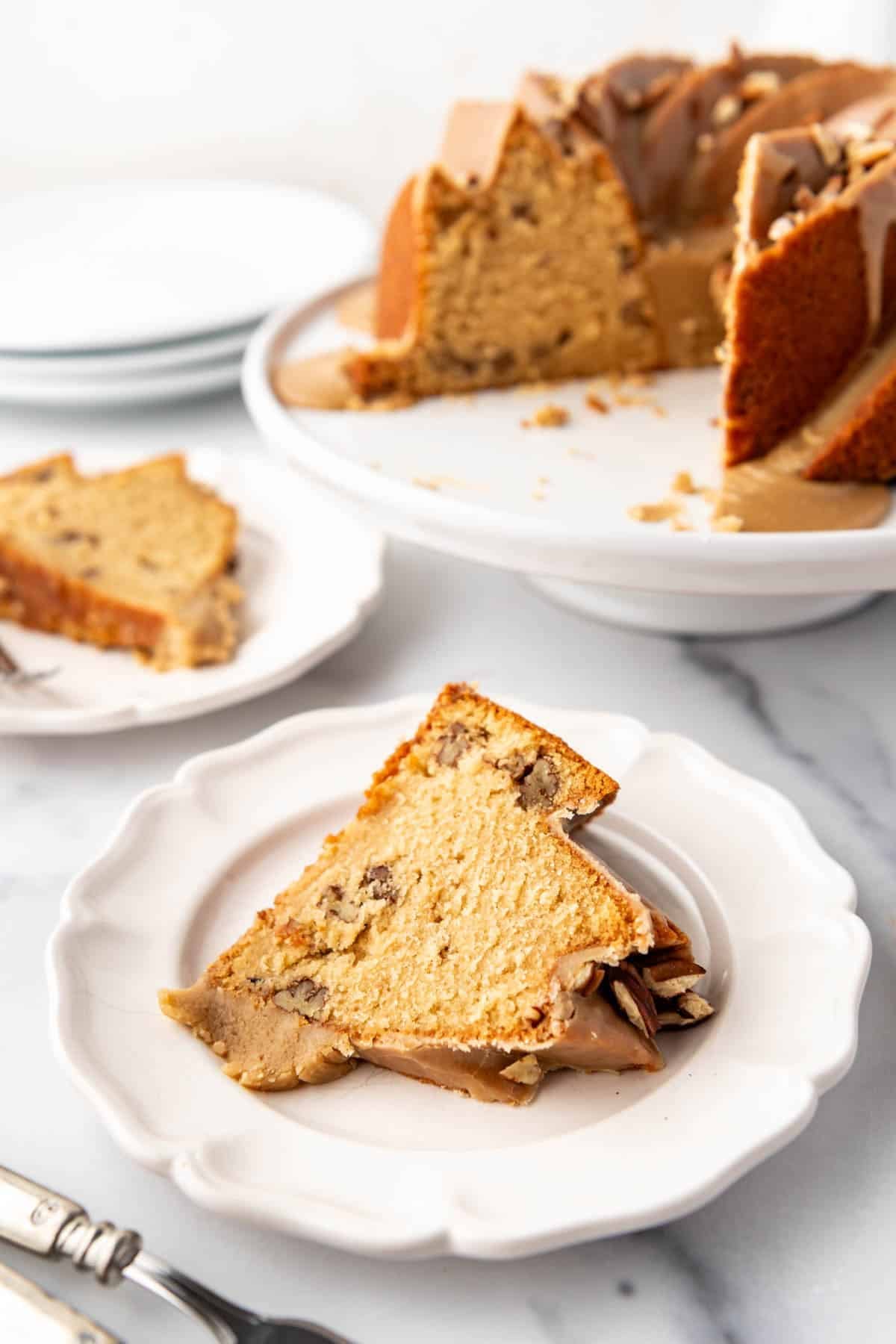 A slice of bundt cake with pecans on a white dessert plate in front of the rest of the cake on a cake stand.
