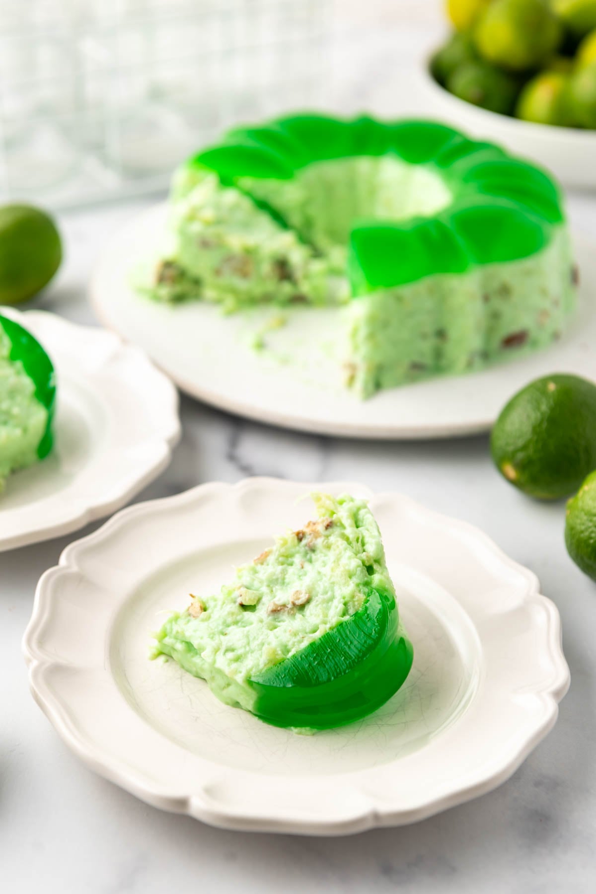 A slice of green lime jell-o on a plate in front of the rest of a lime jell-o mold.