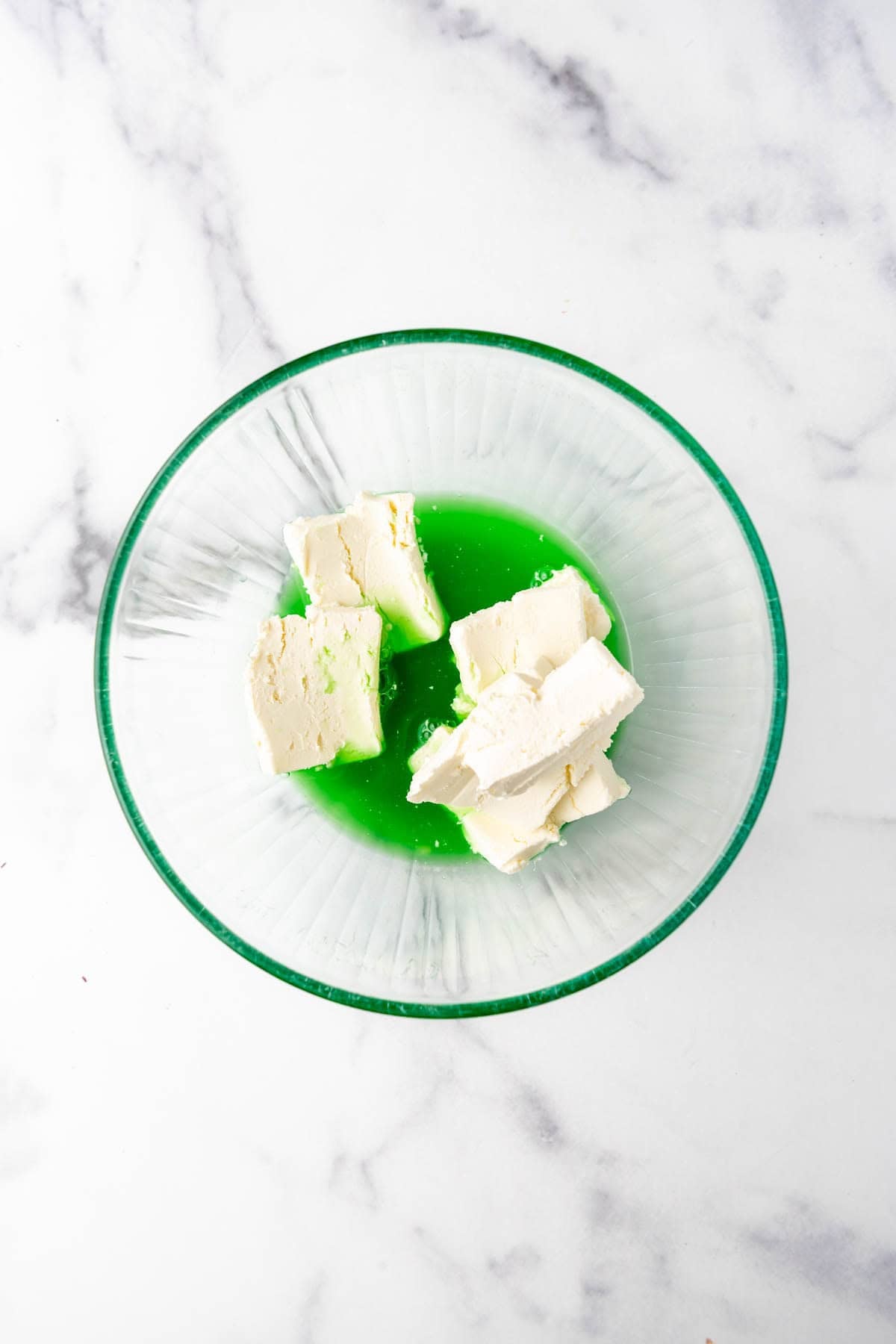 An overhead image of chunks of cream cheese being dissolved into hot lime jello.