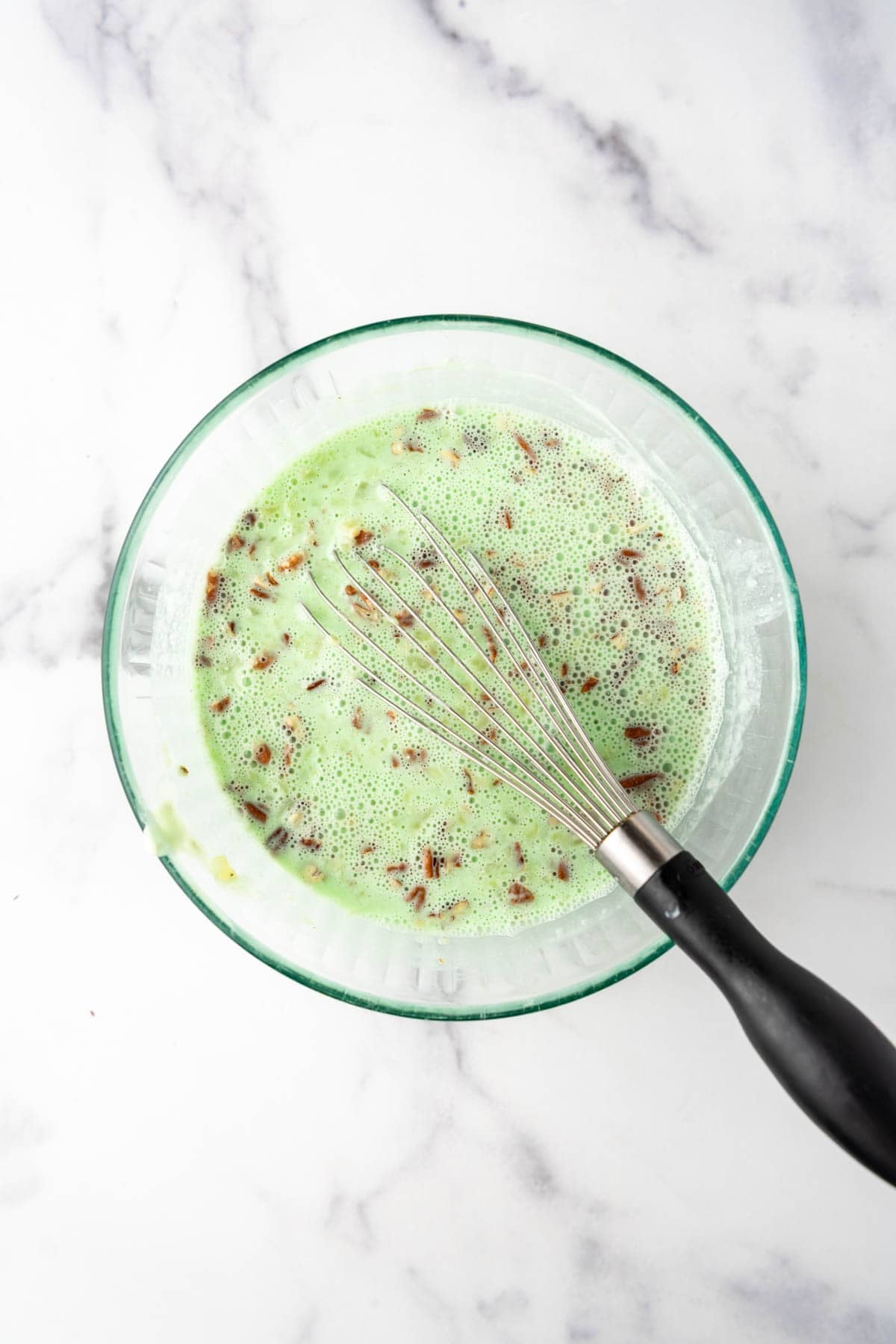 A whisk in a bowl of green jello with chopped pecans and crushed pineapple.