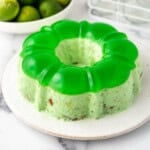A molded lime green jell-o on a white plate with a bowl of limes behind it.
