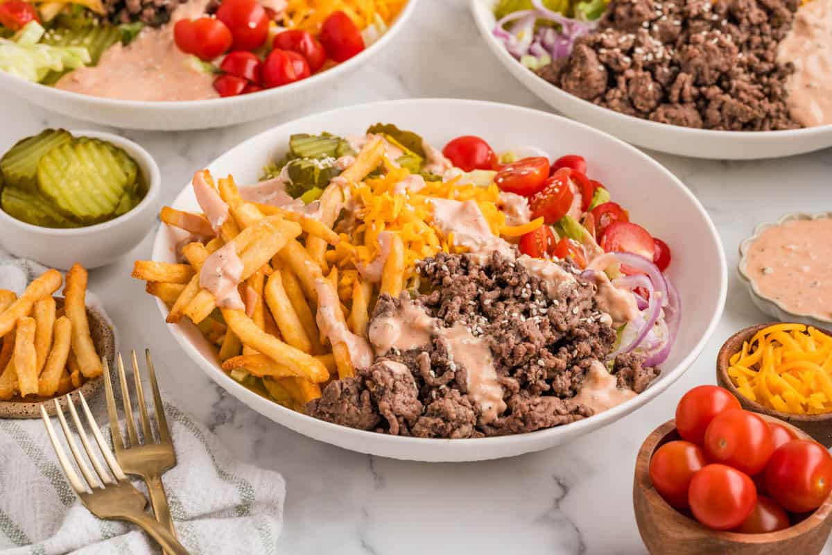 A side angle of a burger bowl surrounded by grated cheese, pickles, tomatoes, and fries.