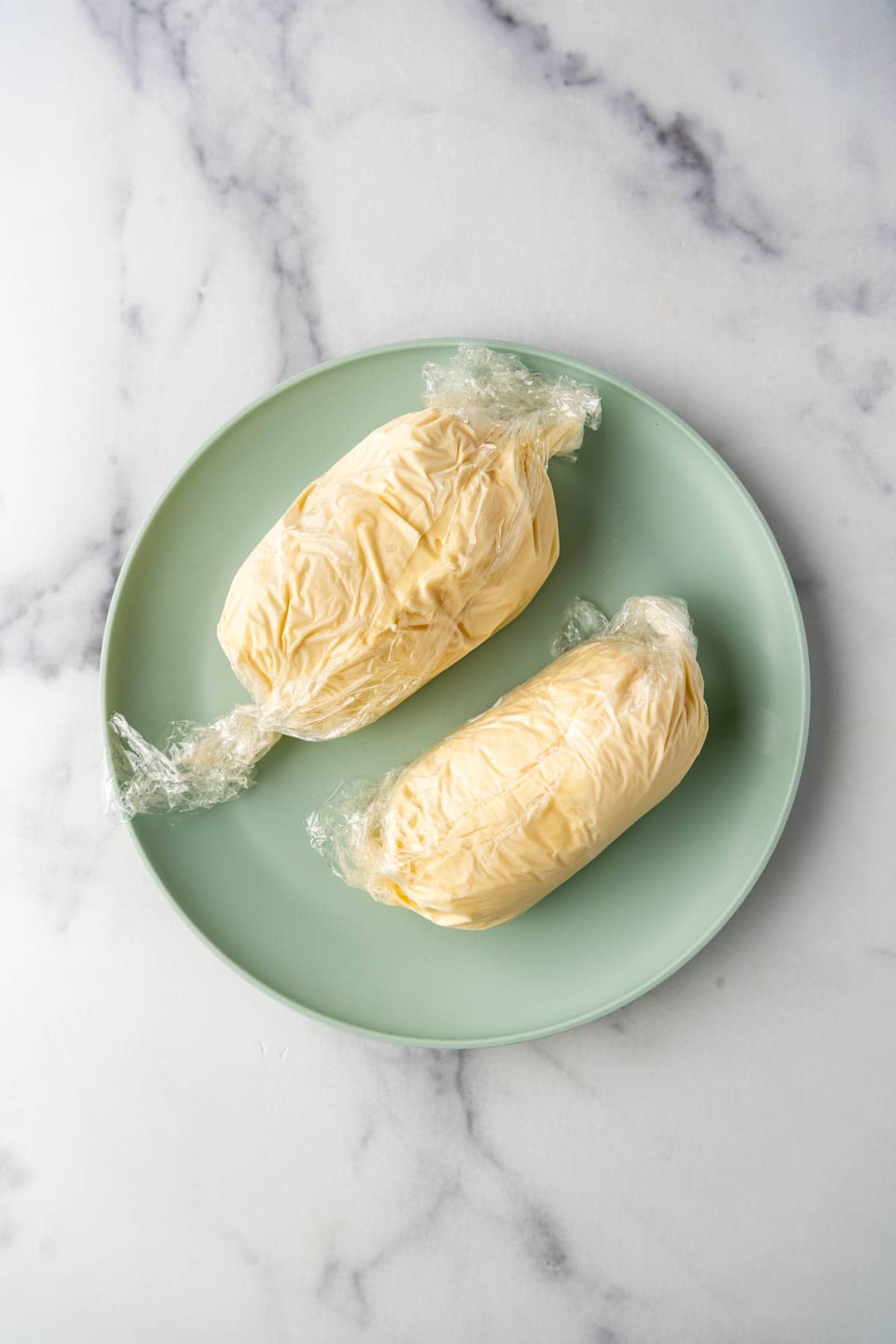 Two oval-shaped mounds of vanilla ice cream wrapped in plastic wrap on a pale green plate.