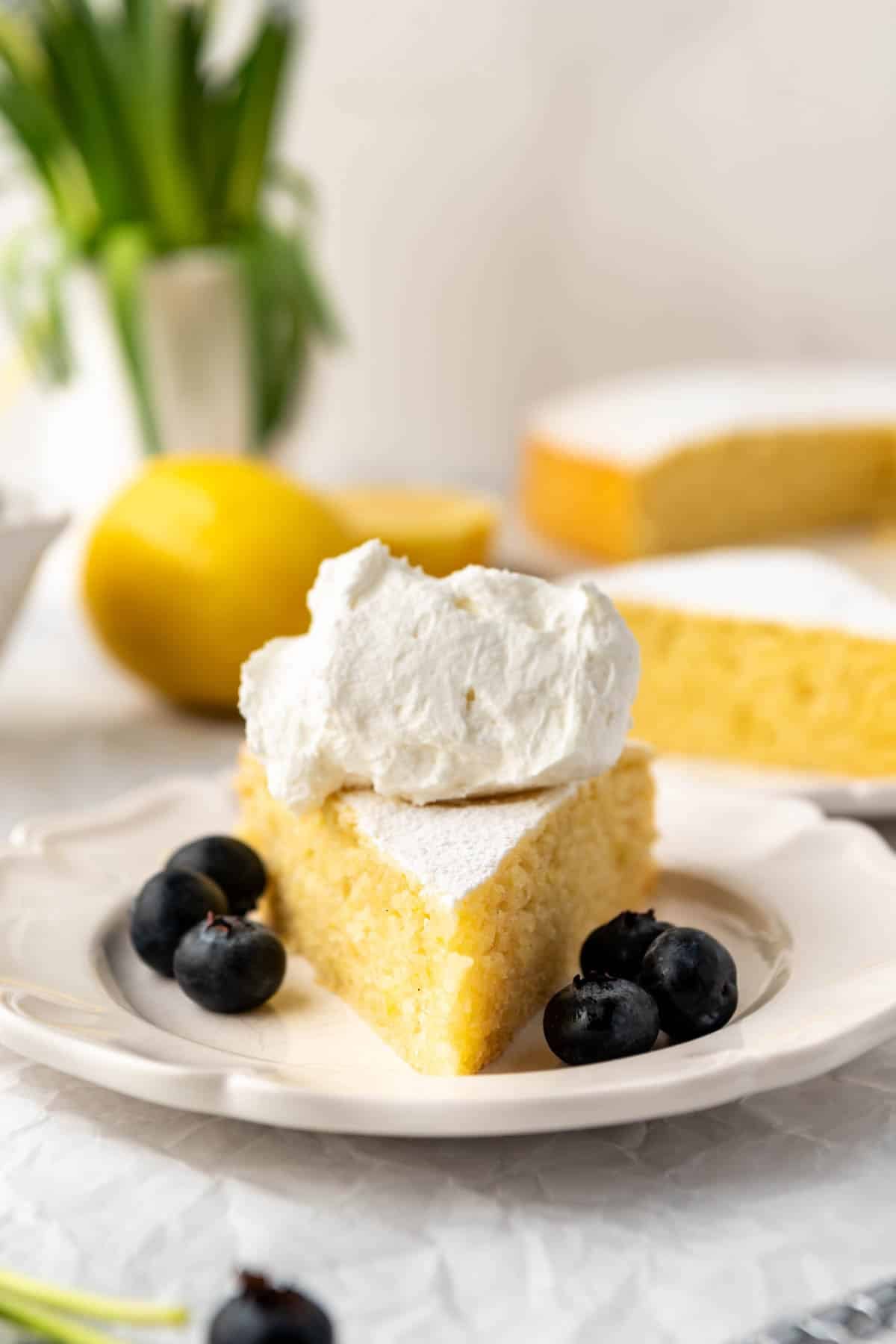 A slice of lemon olive oil cake on a plate with whipped cream on top and blueberries on the side.