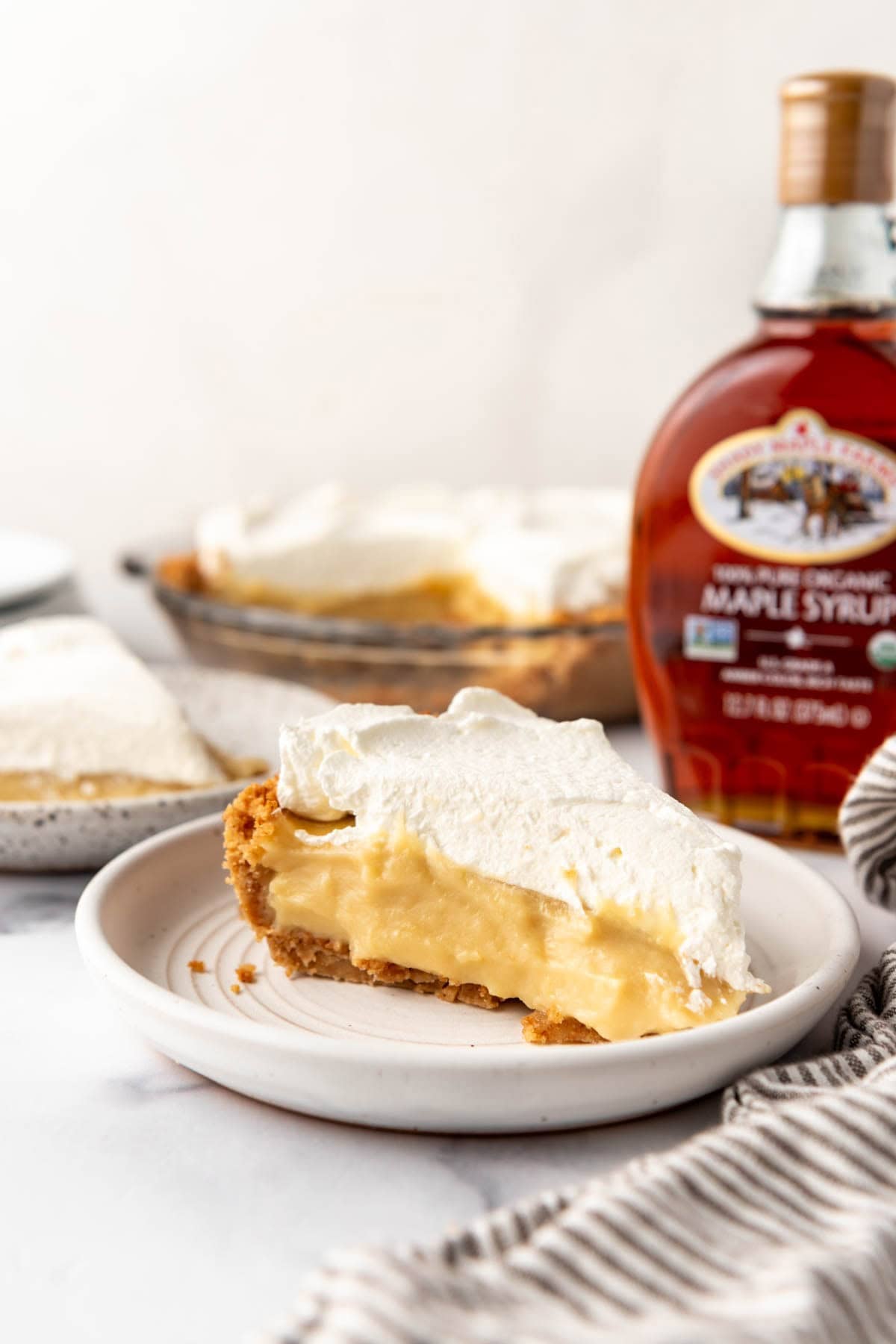 A side image of a slice of maple cream pie on a white plate with a bottle of real maple syrup behind it.