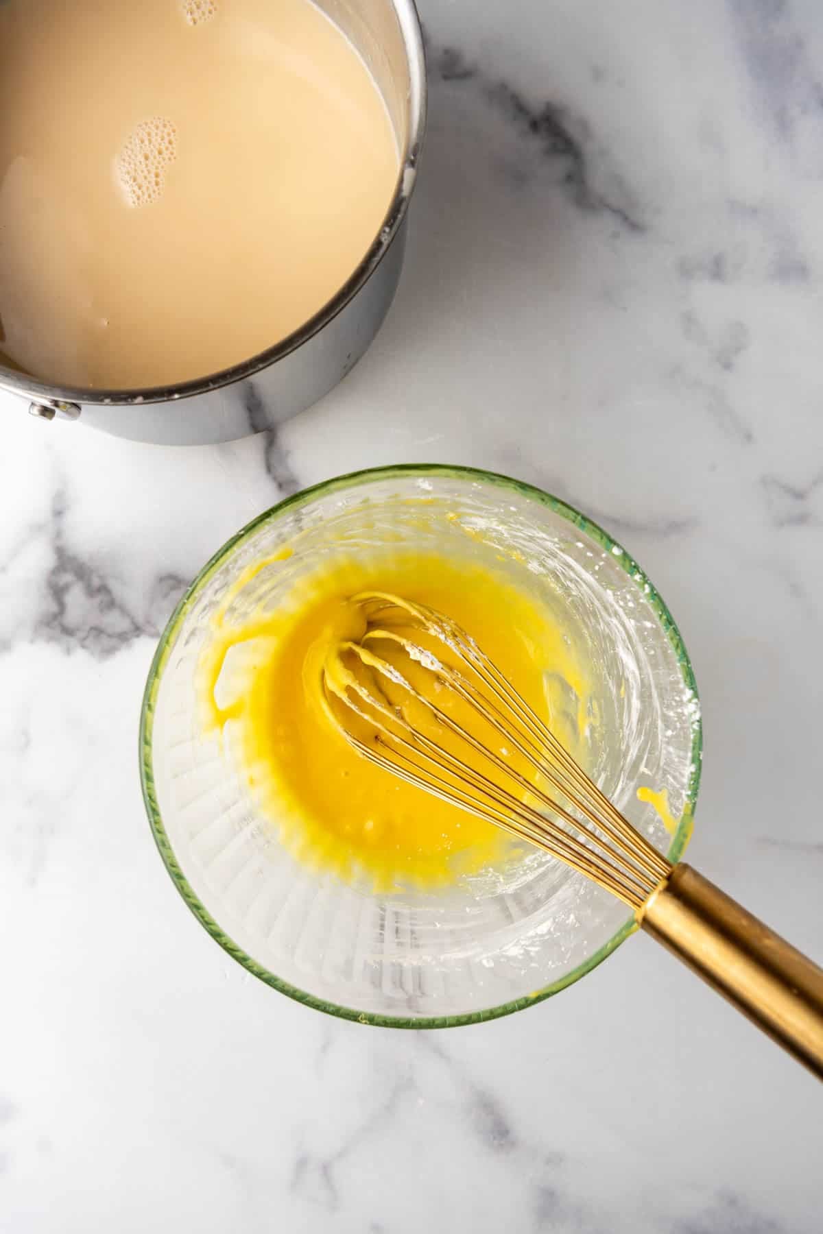 An overhead image of sugar and egg yolks with a whisk in a bowl next to a pan of hot cream and maple syrup mixture.