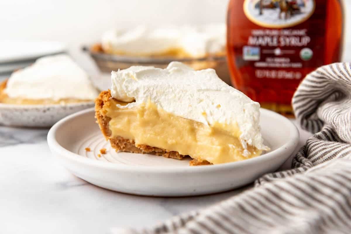 A side image of a slice of maple cream pie on a white plate with a bottle of real maple syrup behind it.