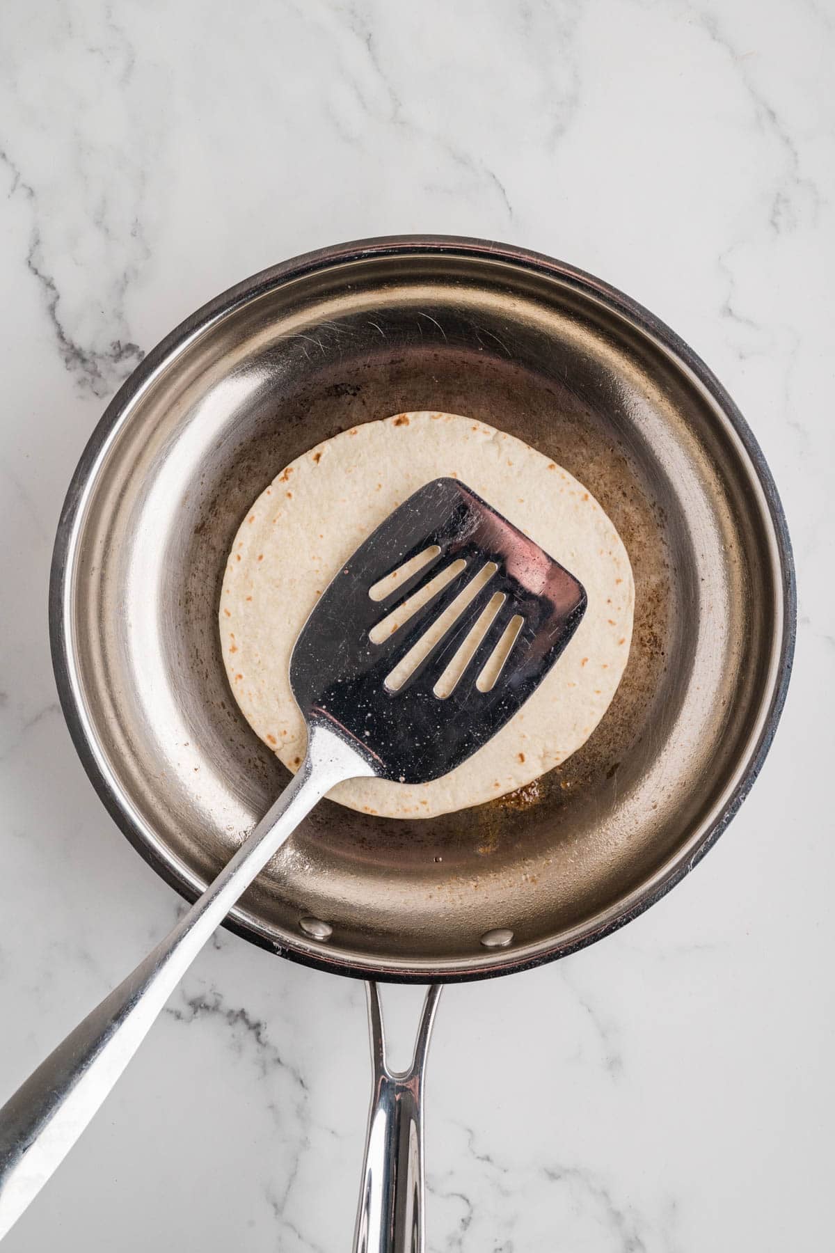 A large spatula being used to flatten a meatball beneath a tortilla in a large skillet.