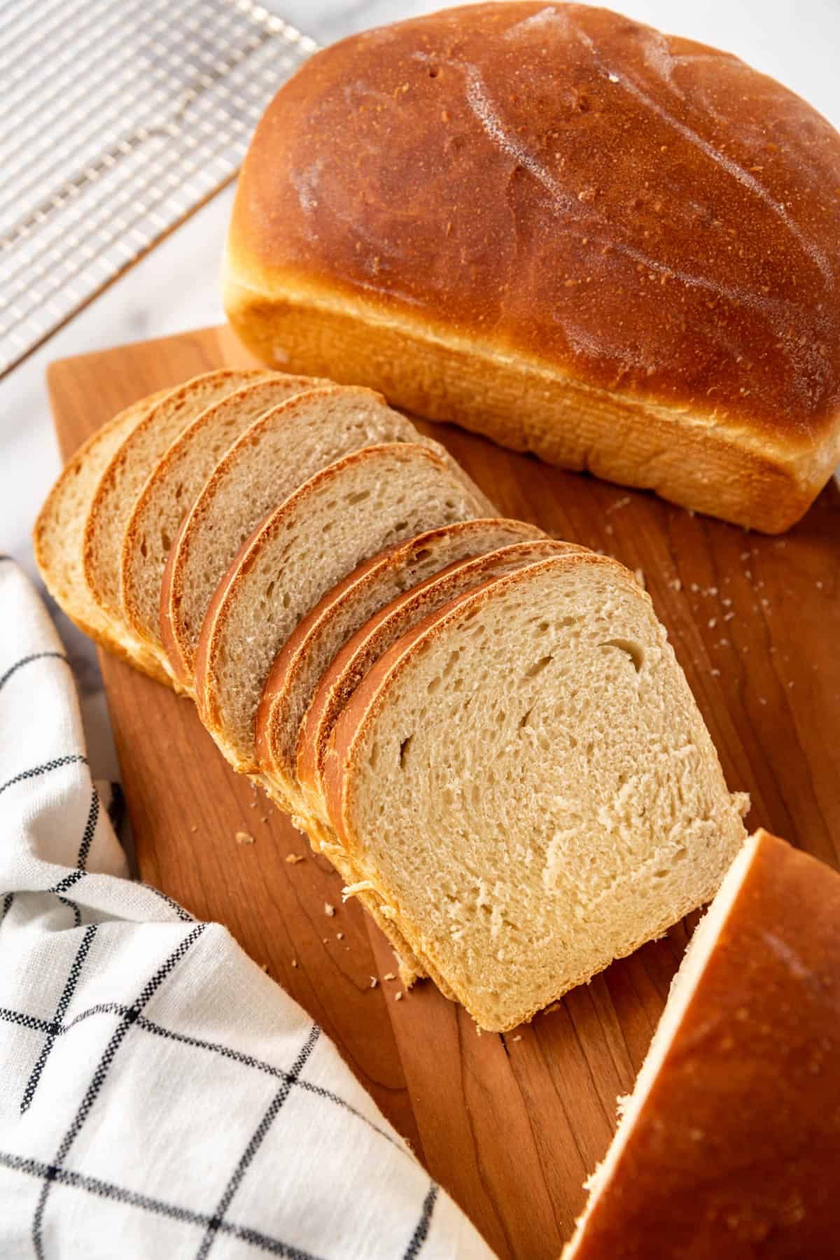 Slices of soft sourdough sandwich bread splayed on a wooden cutting board next to a white checkered linen.