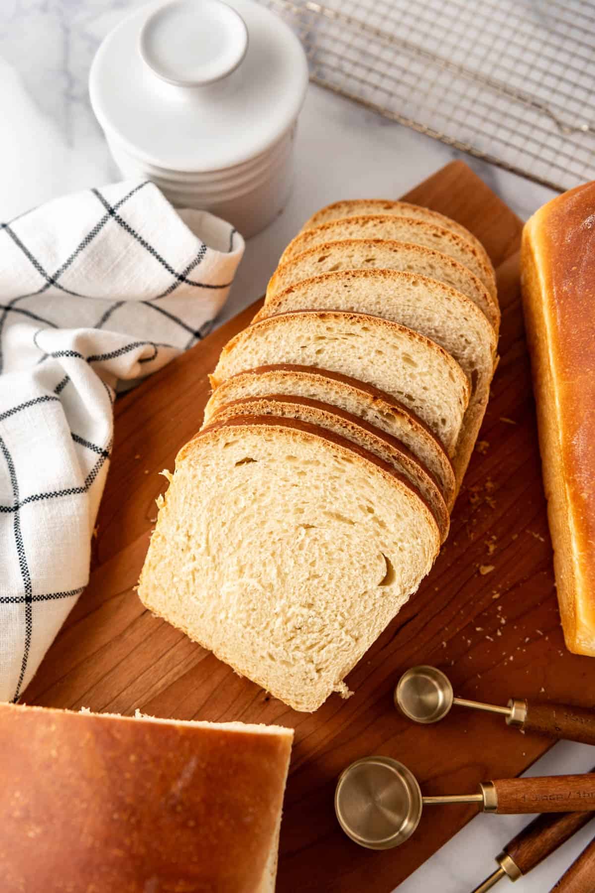 Slices of soft sourdough sandwich bread splayed on a wooden cutting board next to a white checkered linen.