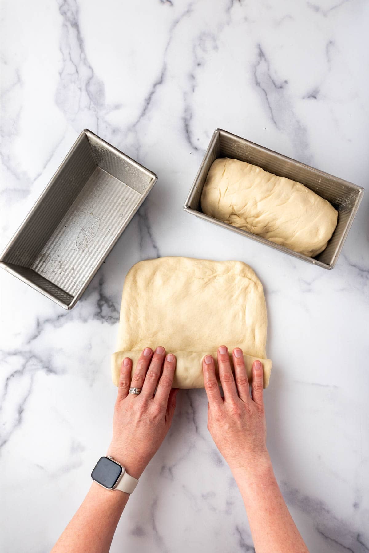 Two hands rolling up a loaf of bread dough with one loaf already in a bread pan.