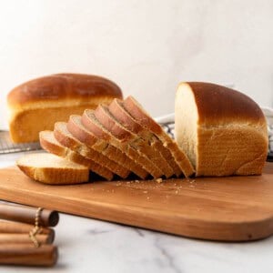 An image of a partially sliced loaf of soft sourdough sandwich bread on a wooden cutting board.
