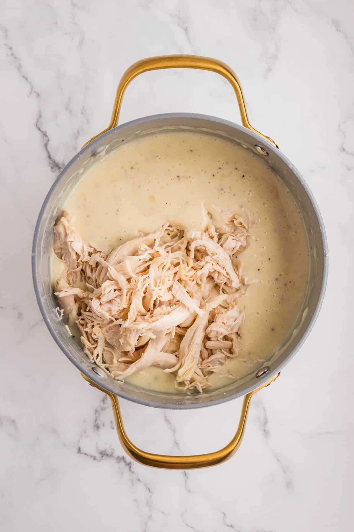 Adding shredded chicken to a pot of creamy sauce.