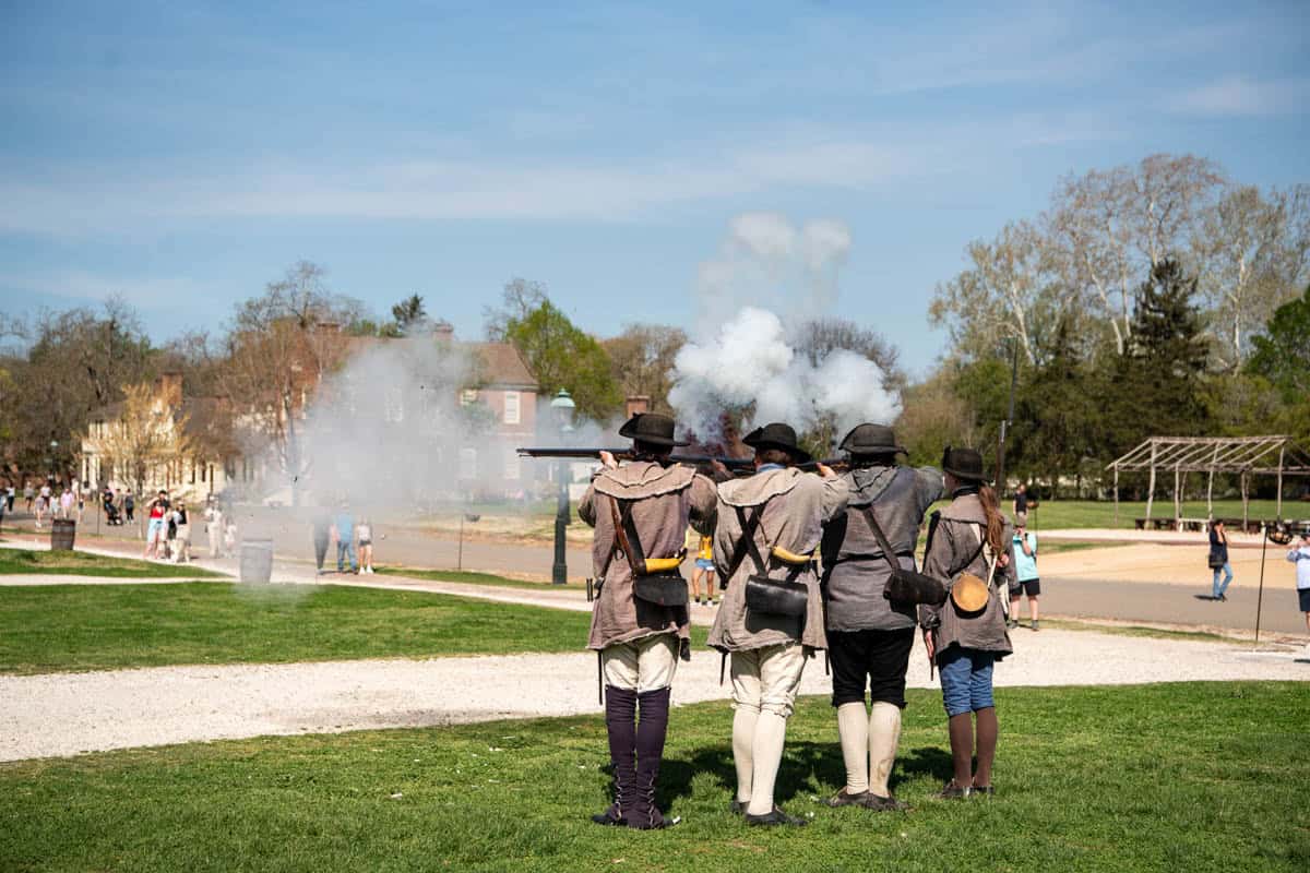 Four revolutionary war soldier living history actors demonstrating firing muskets in Colonial Williamsburg with smoke rising from the just-fired muskets.
