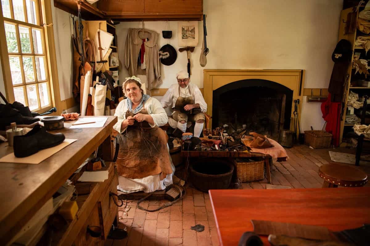 A man and woman in colonial-era clothing working in the leather shop making shoes.