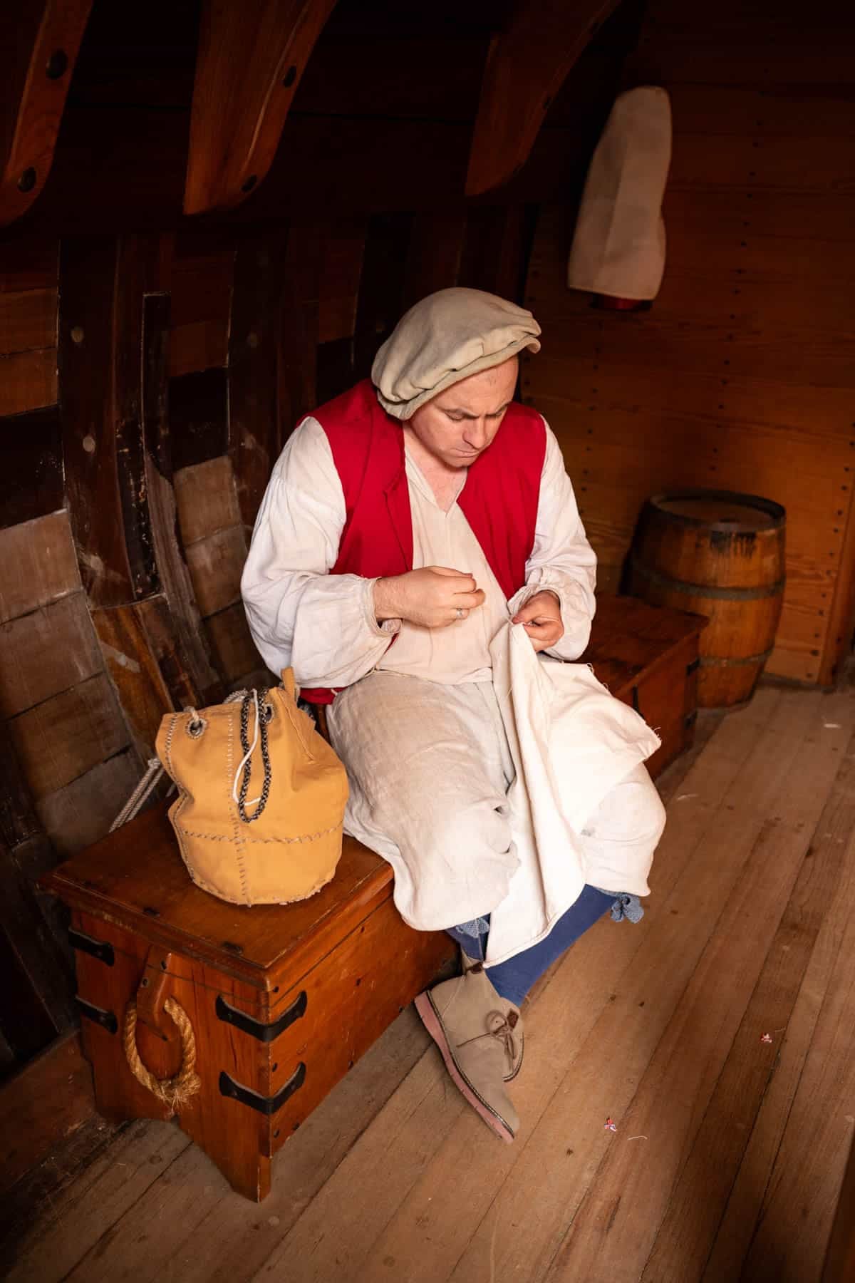 An image of a man in period attire sewing a sail in the hold of a replica sailing ship at Jamestown Settlement colony museum in Virginia.