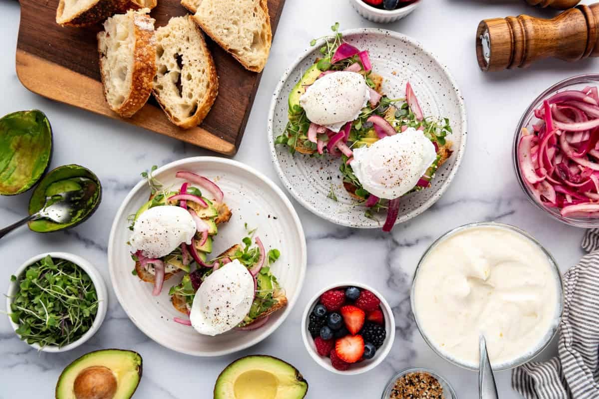An overhead image of two plates of avocado toast with poached eggs, pickled onions, and honey orange ricotta.