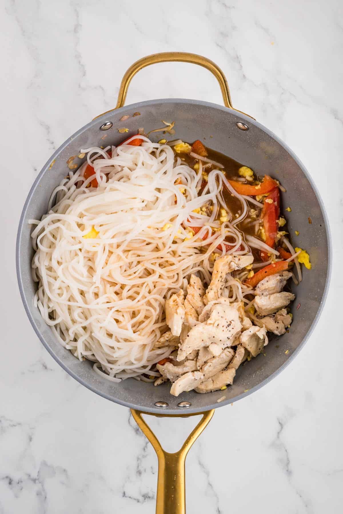 Adding softened rice noodles and cooked chicken to a pan with veggies.