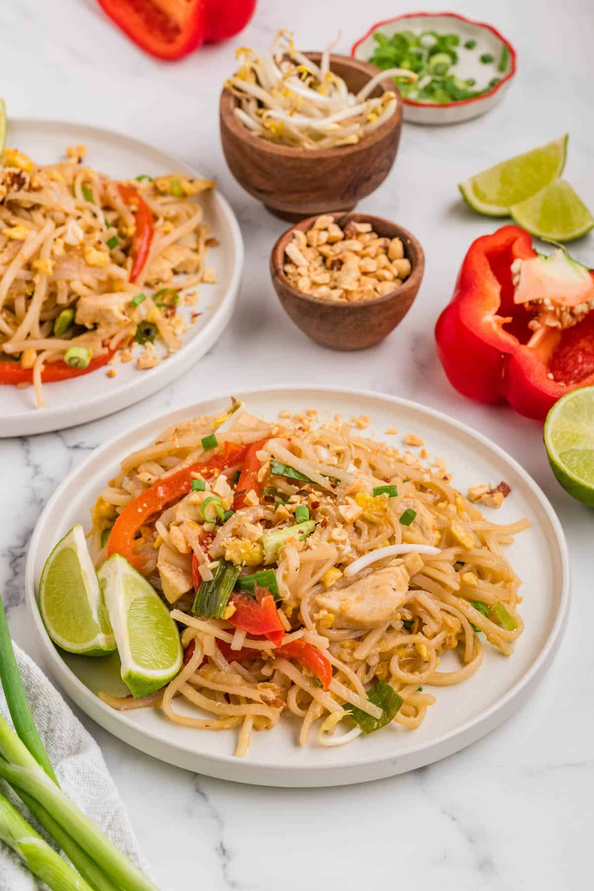 Two plates with servings of pad thai noodles surrounded by veggies.