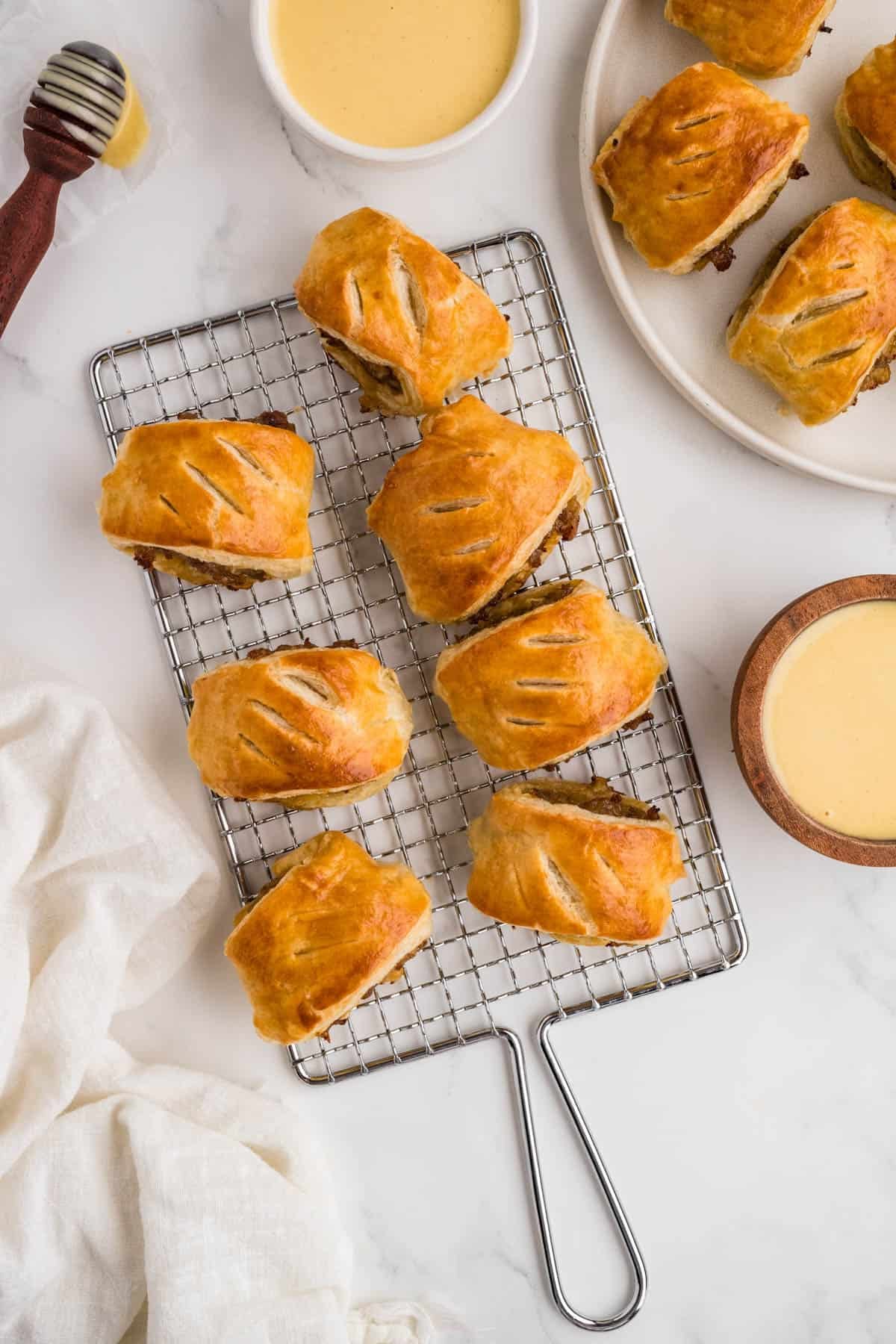 An overhead image of sausage rolls on a wire rack next to a bowl of honey mustard dipping sauce.