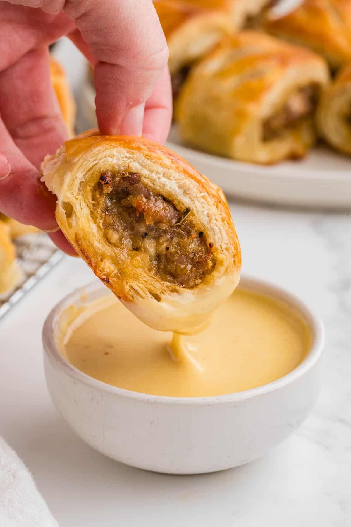 A hand dipping a sausage roll into honey mustard sauce.