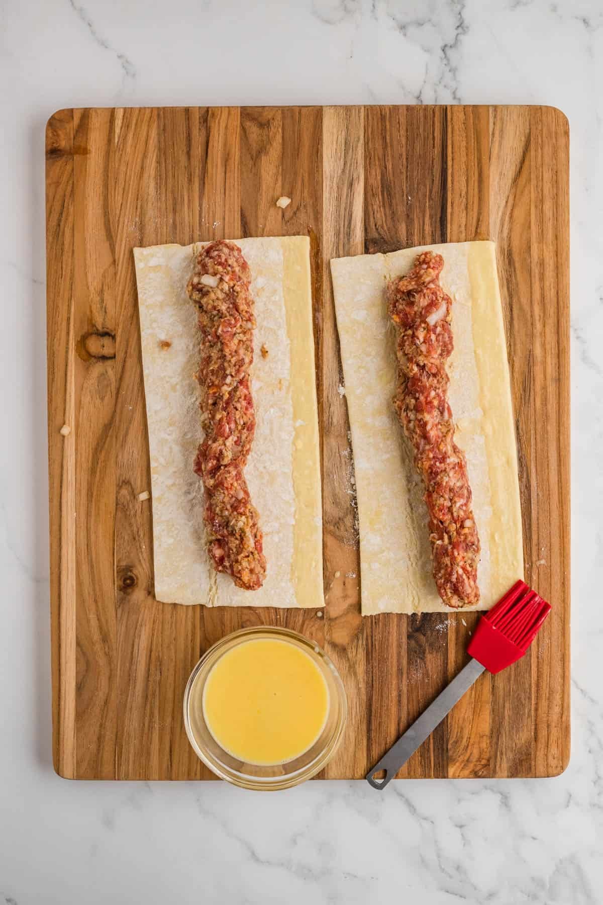 Rows of sausage filling on rectangles of puff pastry next to a bowl of egg wash.