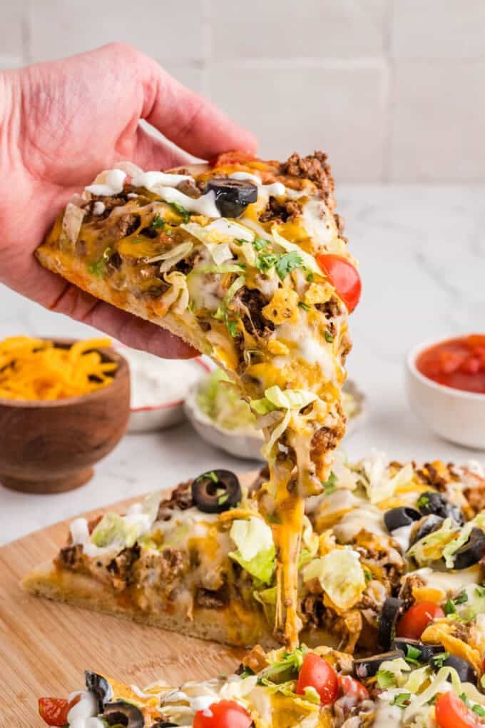 A hand holding a slice of taco pizza.