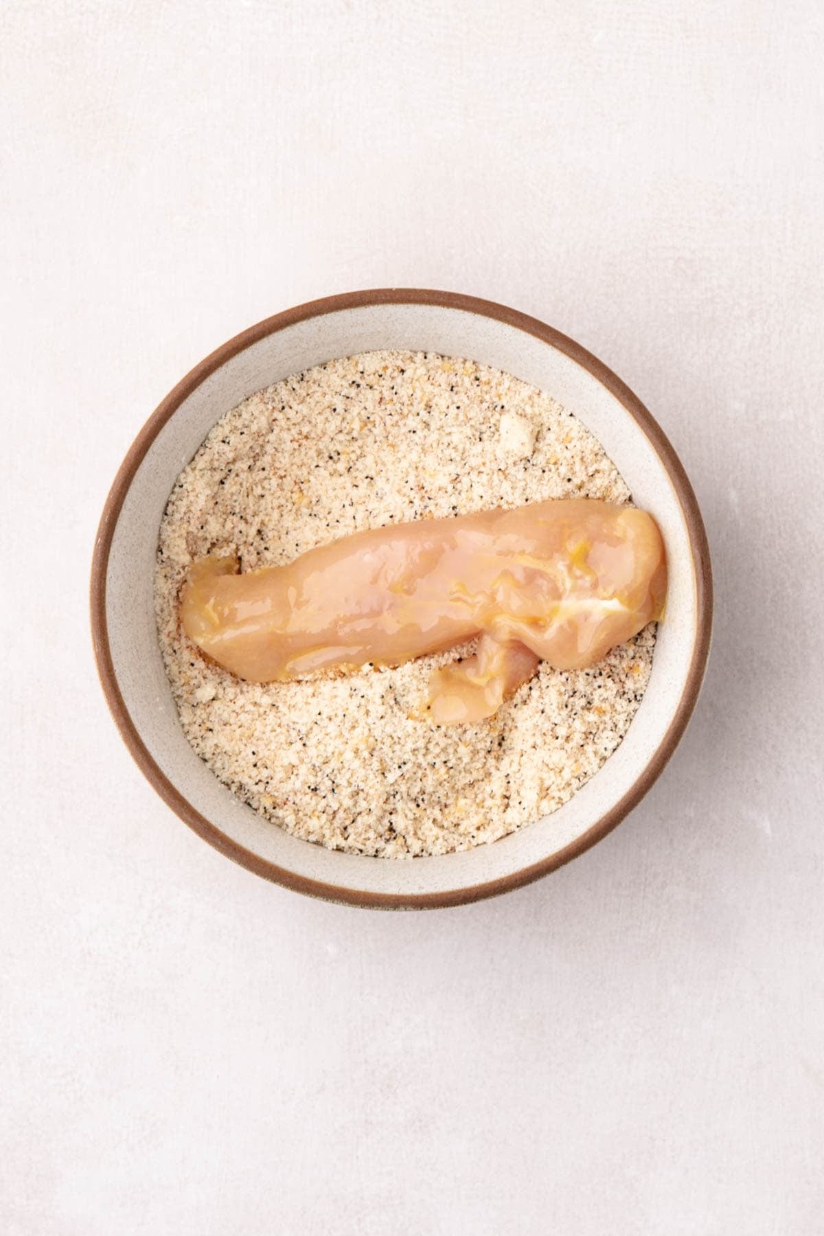 A chicken tender being coated in seasoned breadcrumbs.