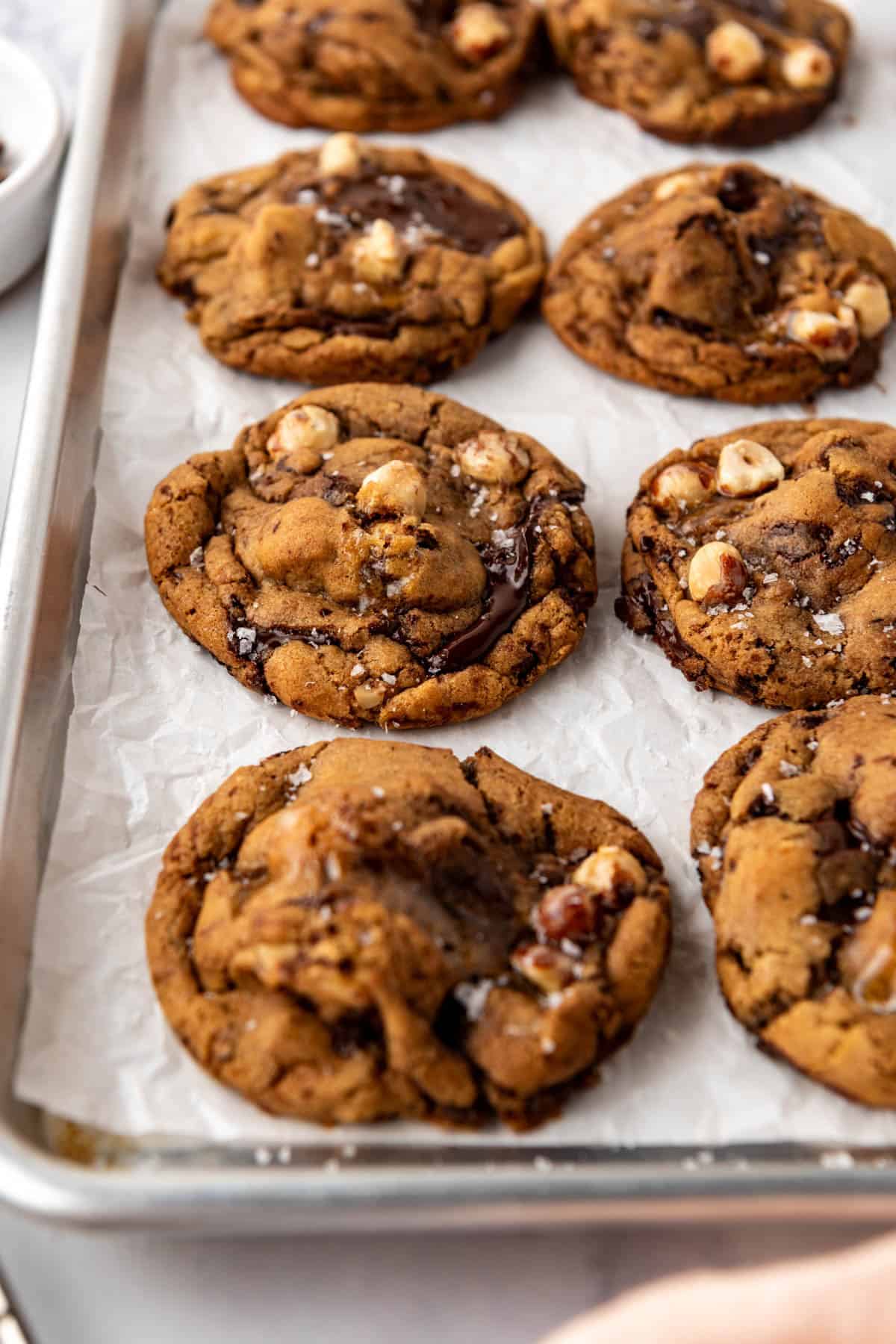 A side angle of cookies on a baking sheet with chocolate puddles and hazelnuts on top.