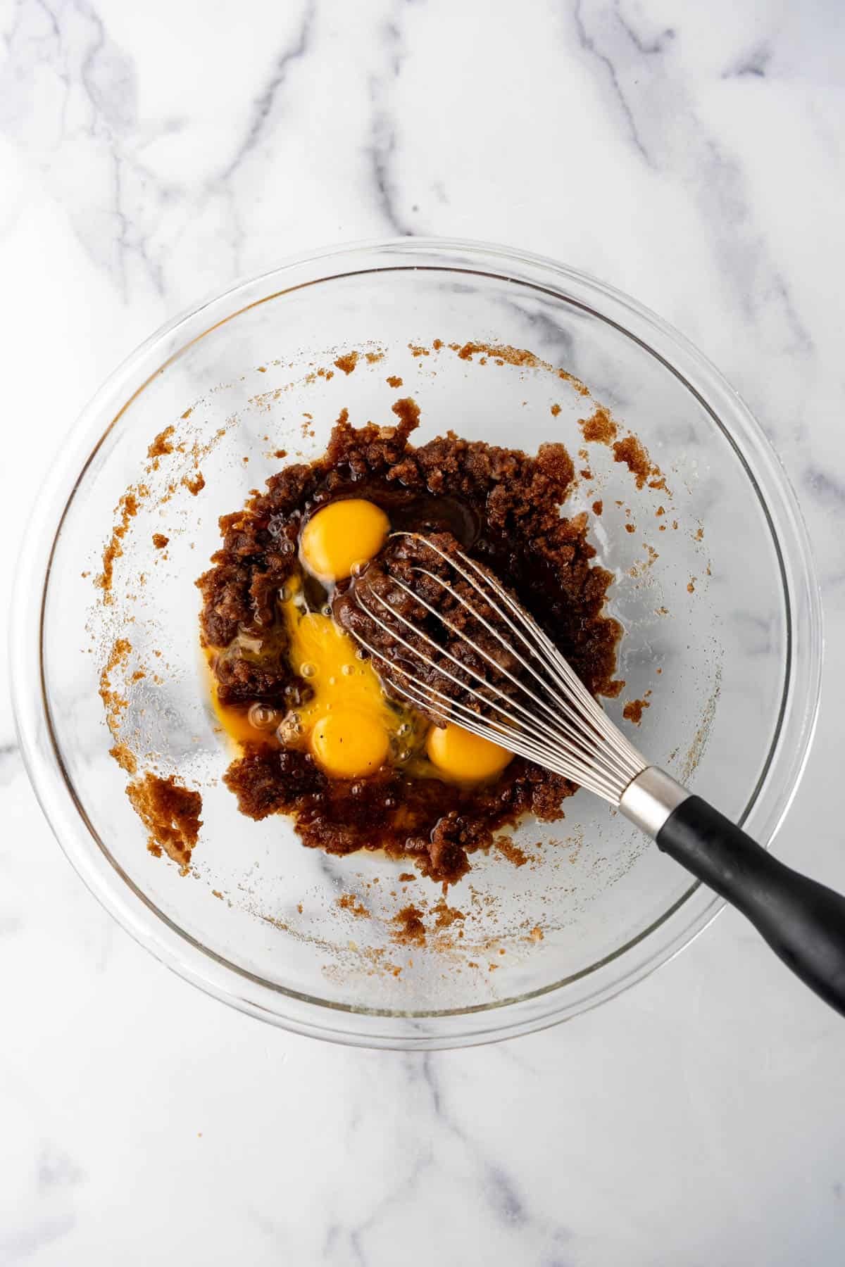 A whisk in a bowl with eggs, egg yolks, vanilla, and creamed brown butter and sugar.