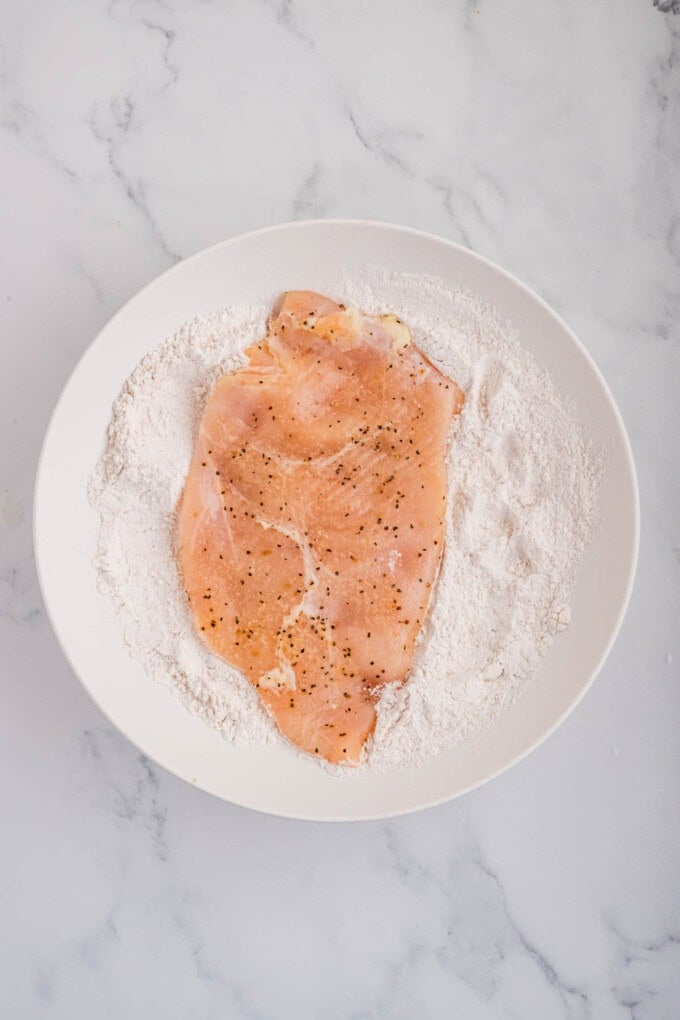 Seasoned chicken breasts being dredged in flour.