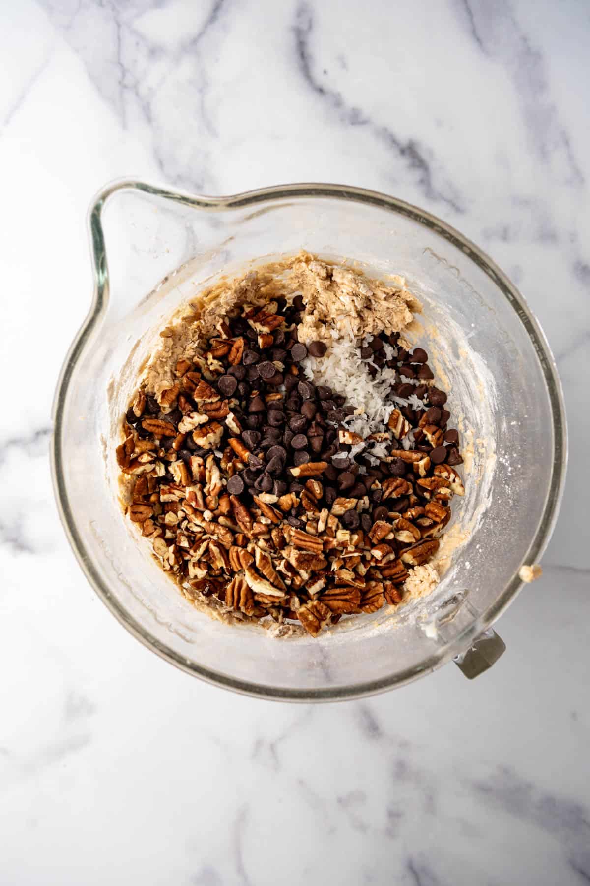 Adding shredded coconut, chocolate chips, and pecans to cookie dough in a glass mixing bowl.