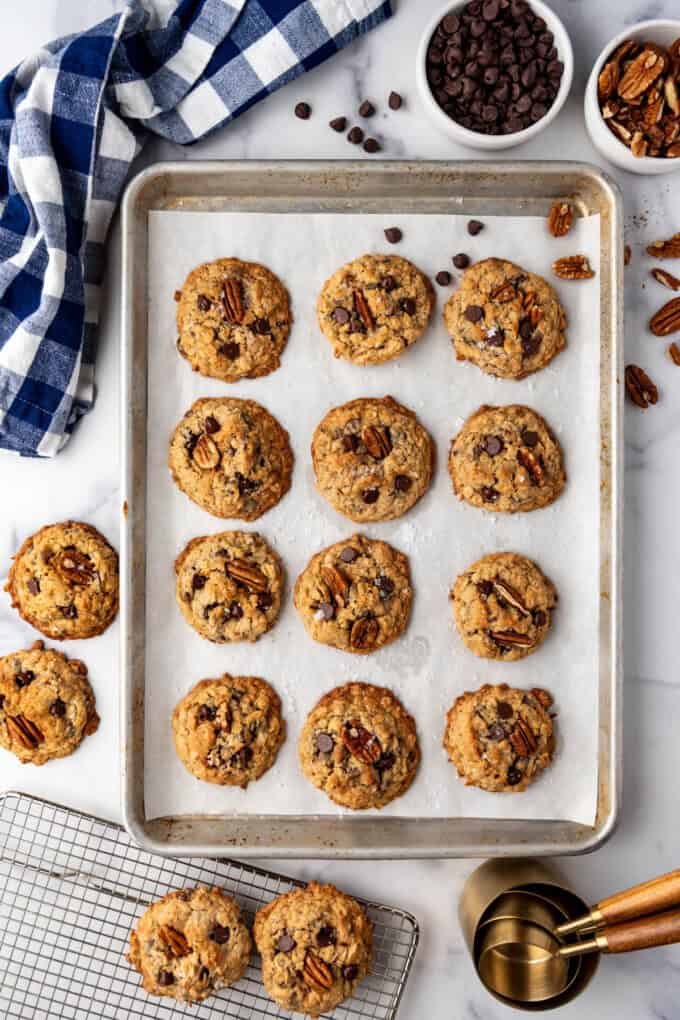 A baking sheet of cowboy cookies surrounded by chocolate chips, pecans, and a blue gingham cloth napkin.