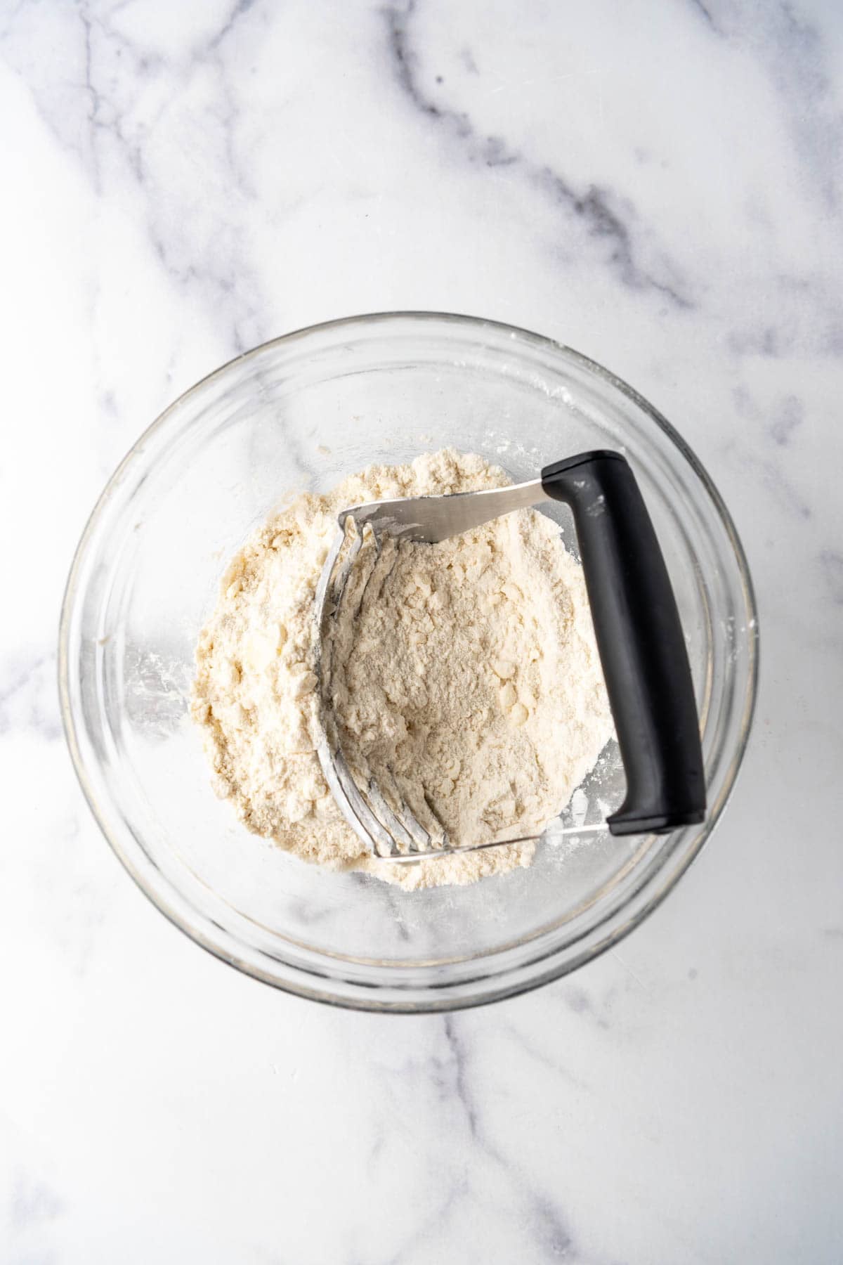 Flour, sugar, and butter that has been mixed into a sandy crumble in a mixing bowl with a pastry cutter.