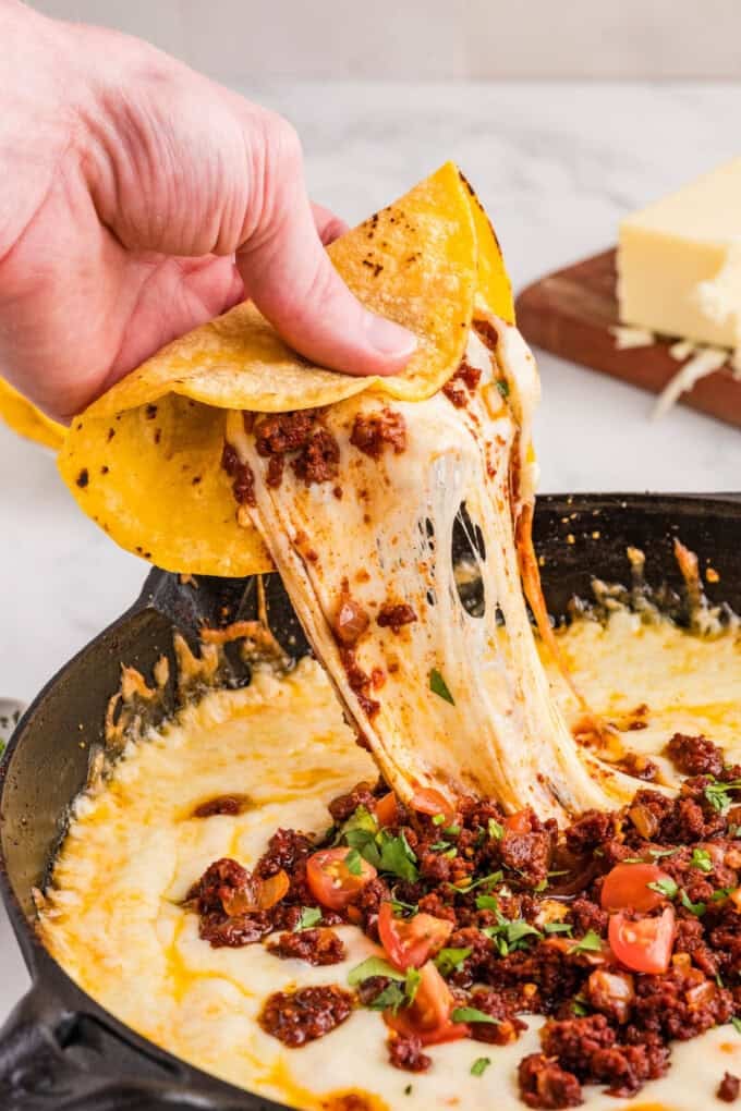 A hand holding a tortilla pulling melted cheese out of a pan of queso fundido.