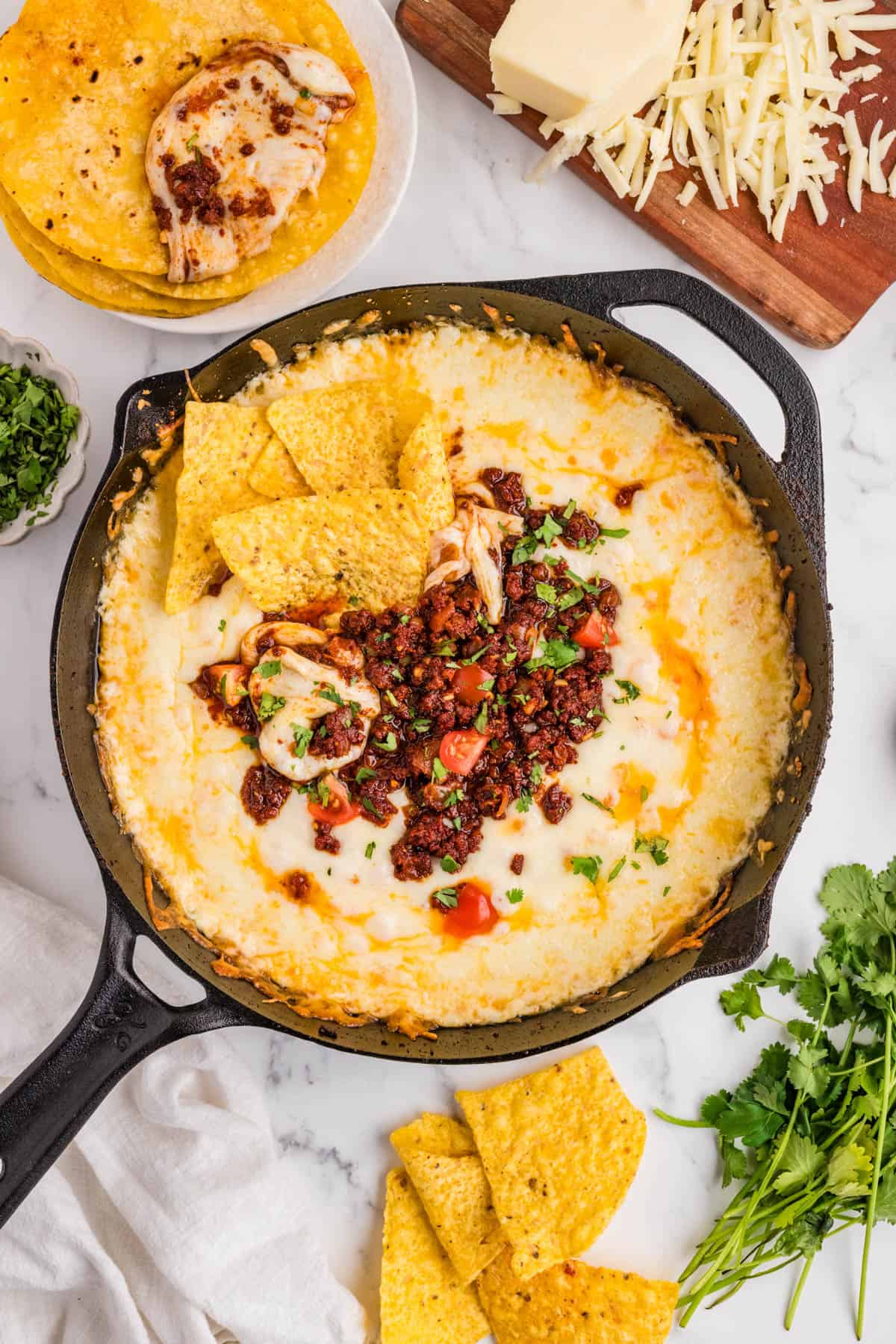 An overhead image of a skillet of queso fundido next to tortilla chips, cilantro, and shredded cheese.