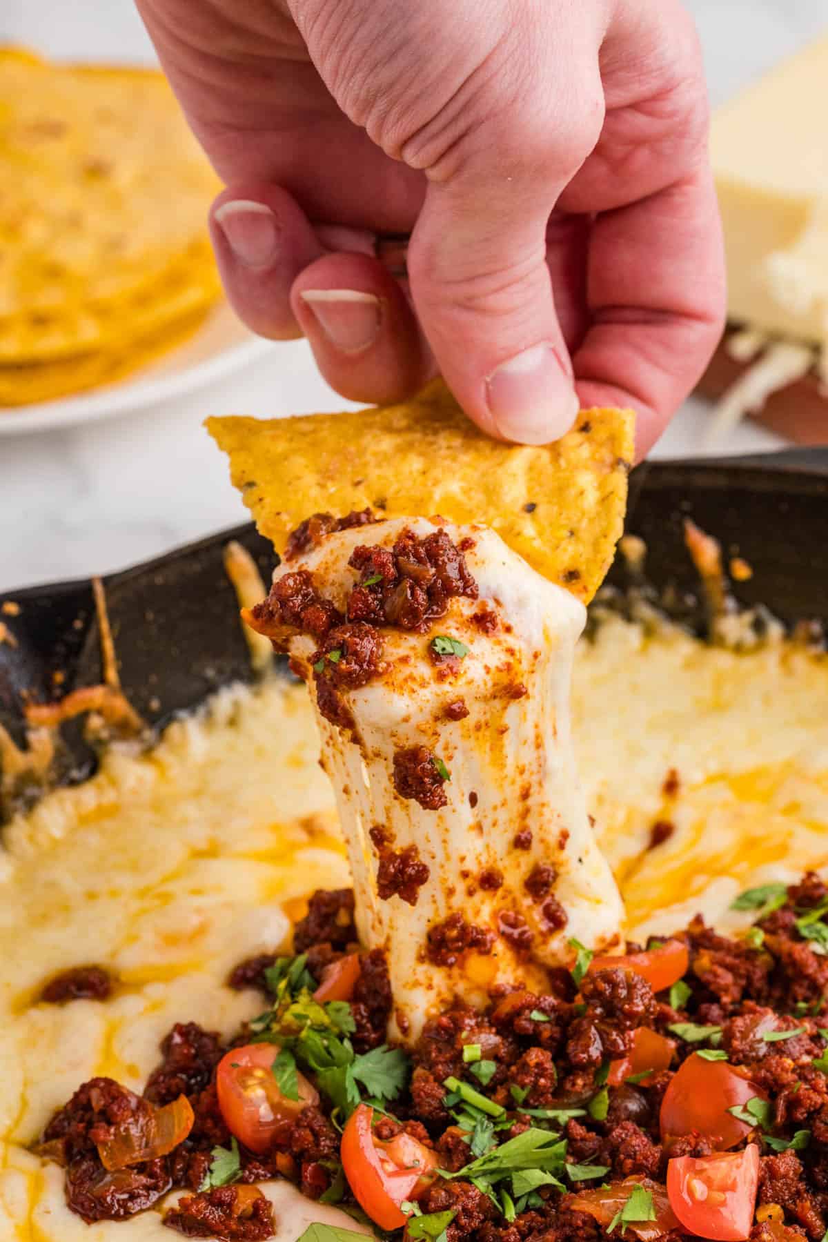 A hand lifting melted cheese and chorizo out of a pan on a tortilla chip.