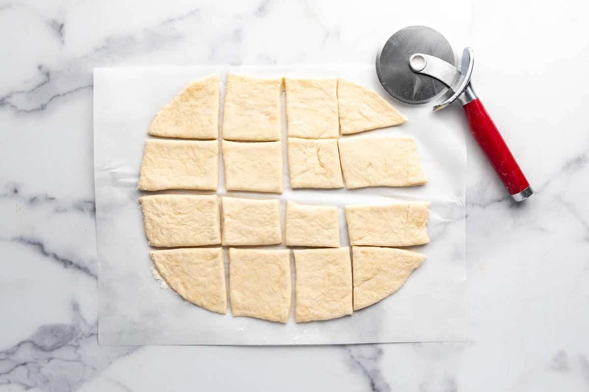 Sopapilla dough rolled out and cut into squares with a pizza cutter nearby.