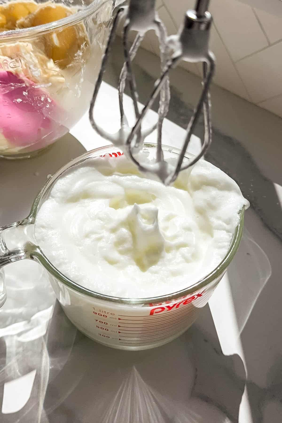 Electric hand mixer beaters being lifted out of a bowl of egg whites whipped to stiff peaks.