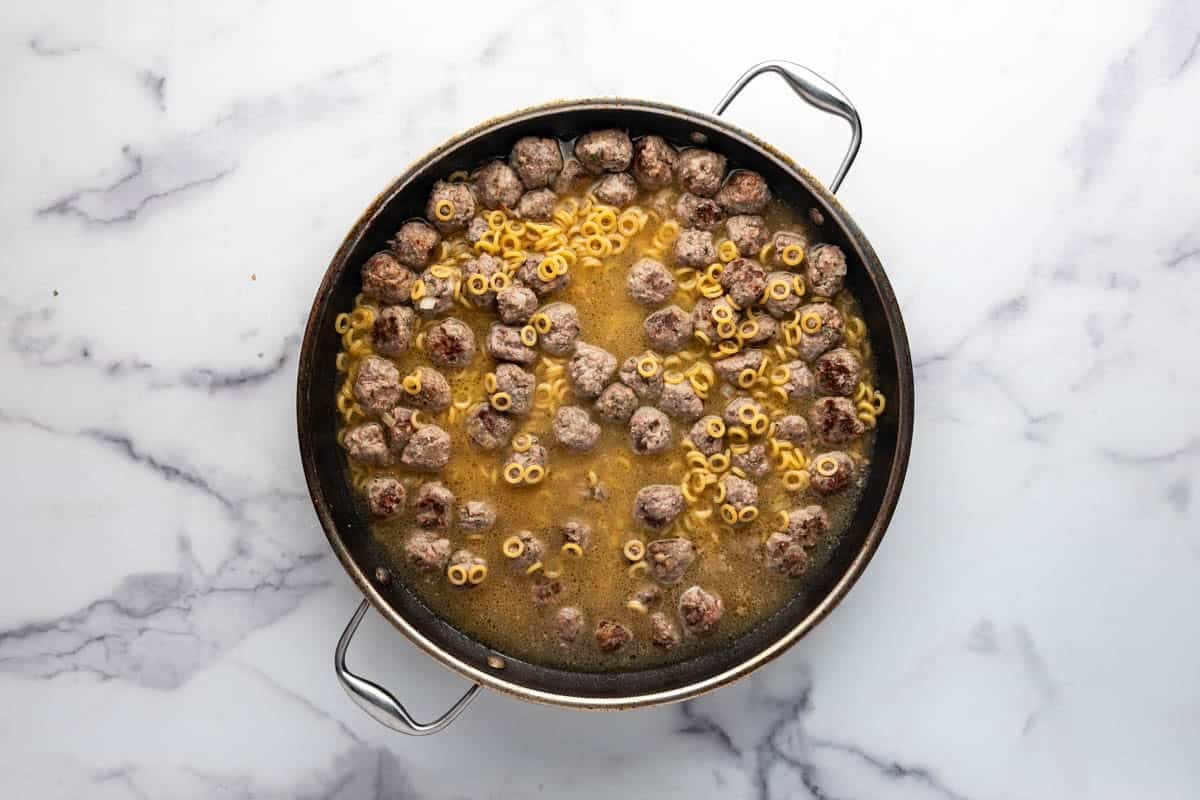 A large skillet with mini meatballs, chicken broth, and pasta.