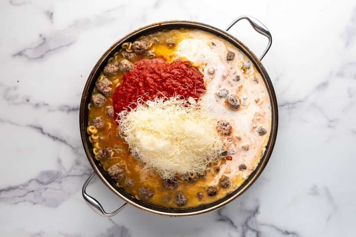 An overhead image of freshly grated parmesan cheese, marinara, and cream being added to a pan of meatballs and pasta.