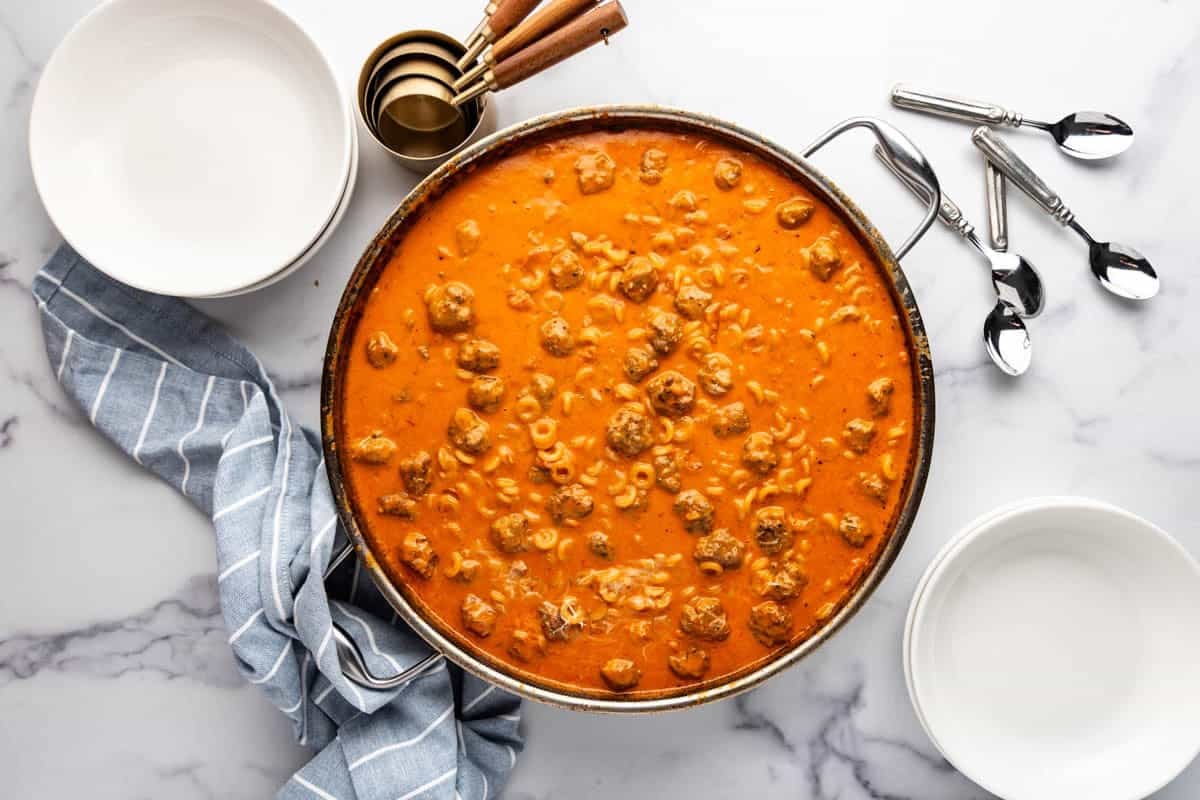 An overhead image of a large pan of homemade spaghettios next to spoons and white bowls.