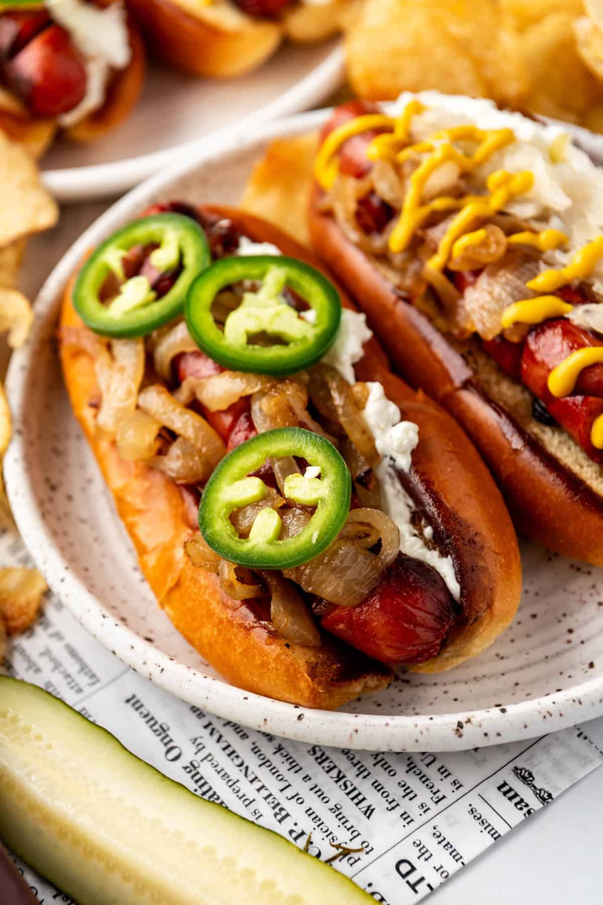An image of Seattle-style hot dogs on a plate with a pickle spear in front and chips in the background.