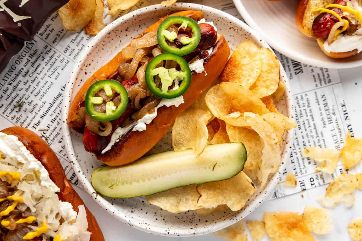 Hot dogs on buns with cream cheese, sliced jalapenos, and caramelized onions  with chips and pickle spears surrounding them.