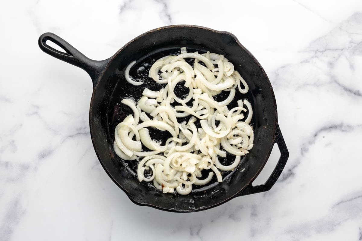 Thinly sliced onions being sauteed in a large cast iron skillet with butter.