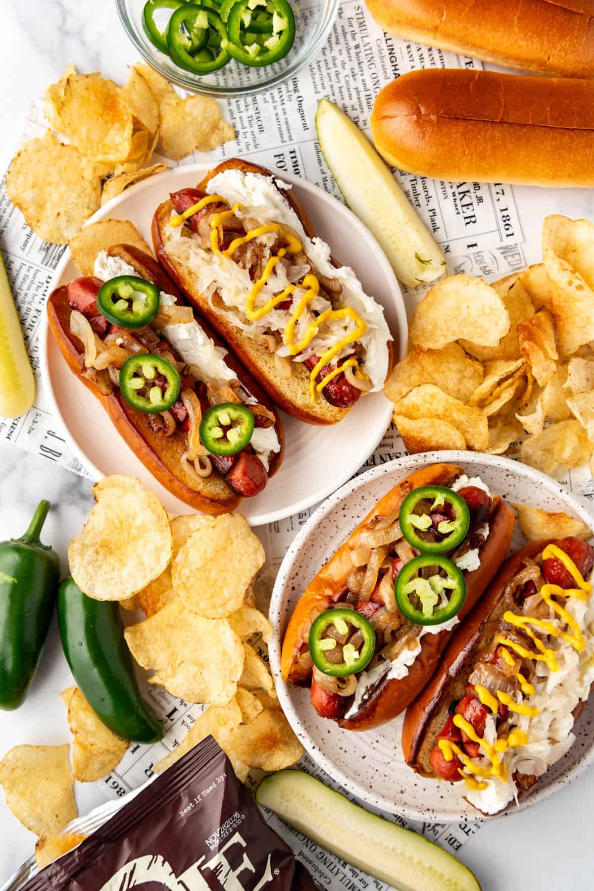An overhead image of Seattle hot dogs on plates with chips, jalapenos, and pickle spears surrounding them.