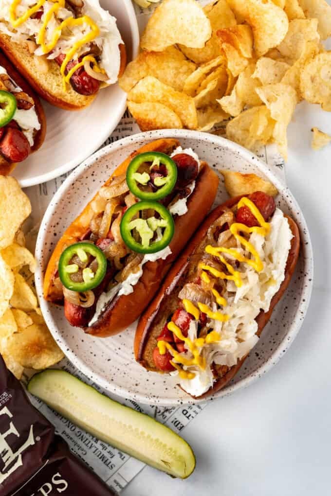 An overhead image of gourmet hot dogs on a plate with pickles and chips nearby.